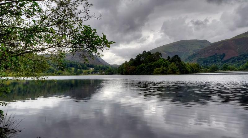 Calm lake with overhanging tree branches in the foreground, surrounded by green hills under a cloudy sky.