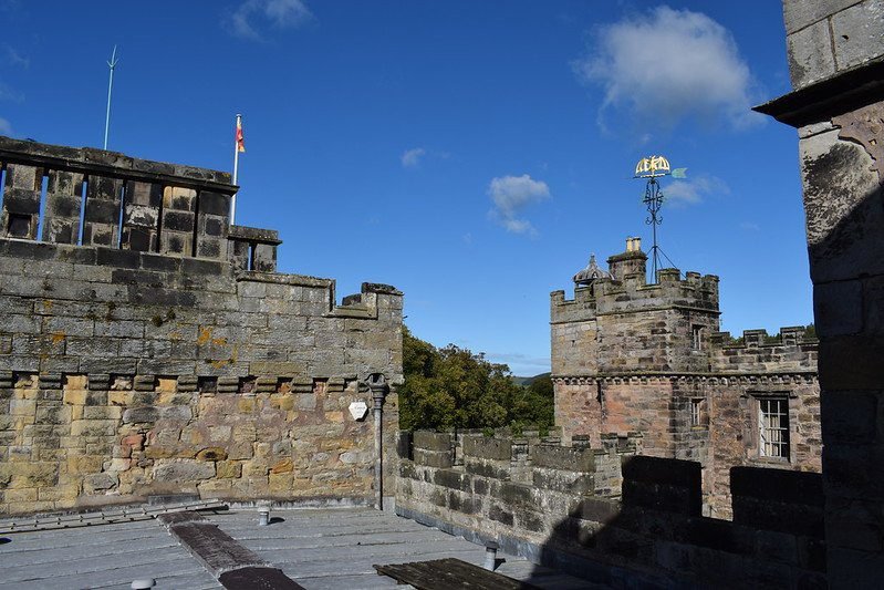 Rooftop view of a stone castle with battlements and a tower featuring a weather vane against a blue sky.