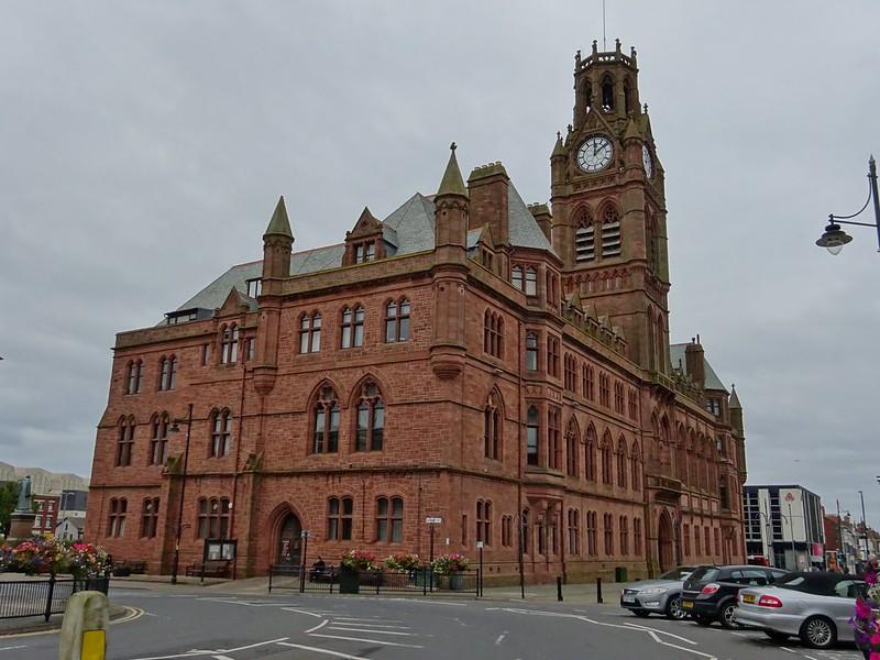 A large red-brick, clock-tower building with Gothic architectural elements, surrounded by a few parked cars, flowers, and a lamppost on an overcast day.