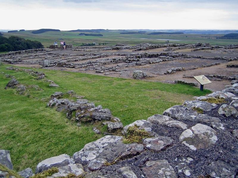 Ruins of a Roman fort with stone foundations and grassy areas against a backdrop of green fields and distant hills.