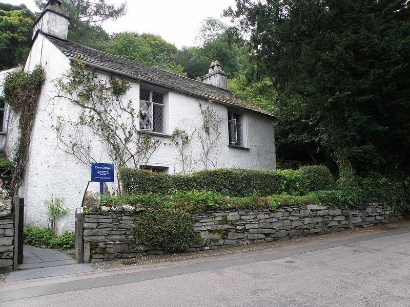 Dove Cottage | White cottage with a grey slate roof, surrounded by greenery and a stone wall along the street.