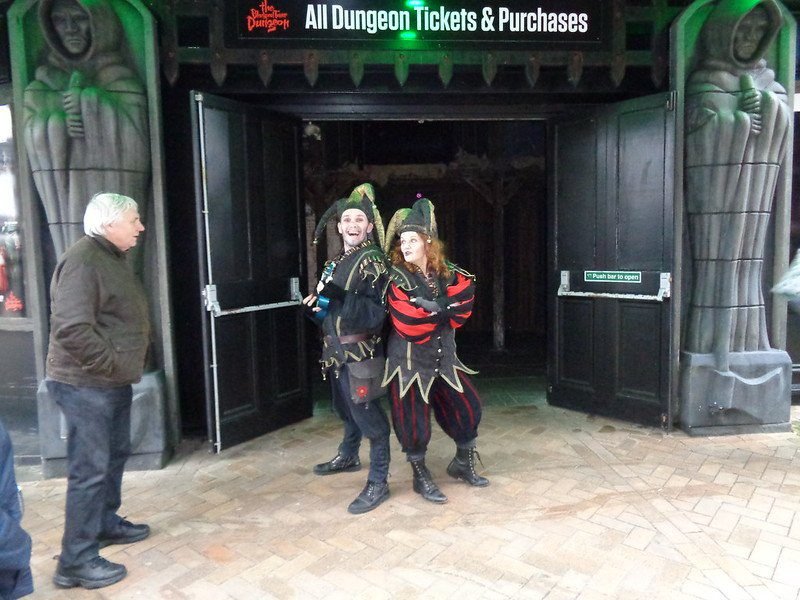 Two performers dressed as jesters stand back-to-back at the entrance of a dungeon attraction, with a man watching them.