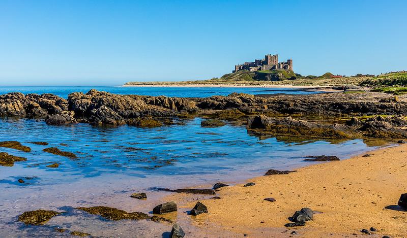 A coastal view with a sandy beach and rocky shoreline, featuring a distant, large castle situated on a hill under a clear blue sky.