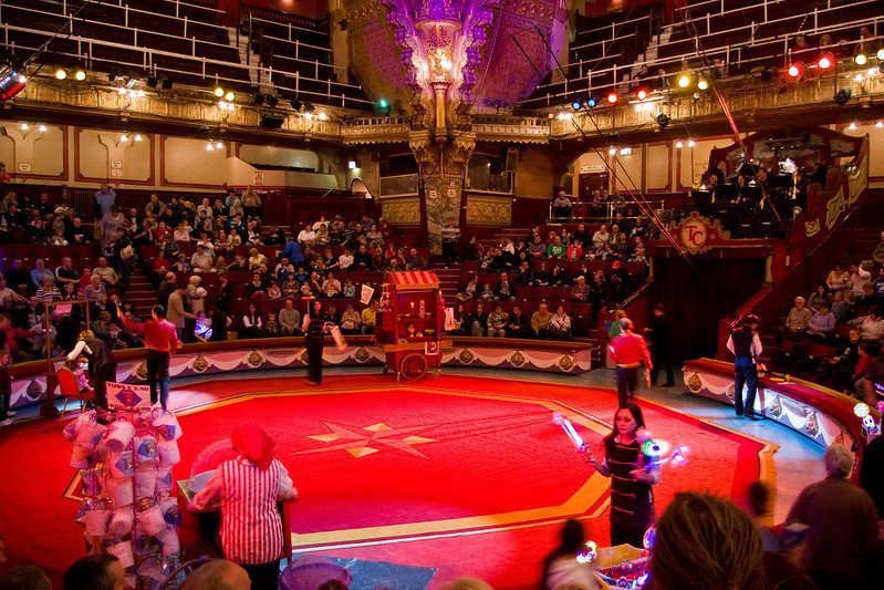 A colourful circus arena with performers and an audience seated in a decorative theatre setting, featuring bright lights and ornate architecture.