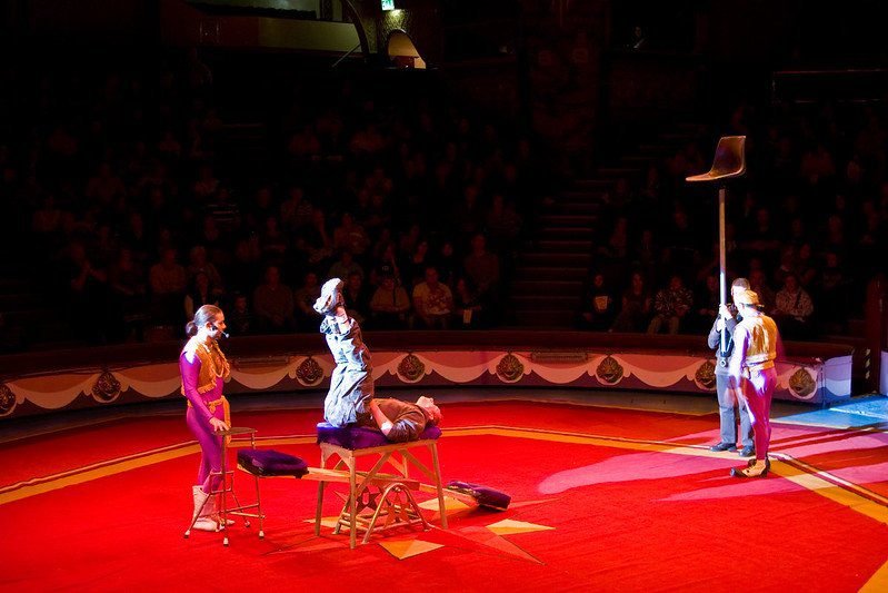 Circus performers on a red stage, two balancing and one holding a long pole with a chair on top.