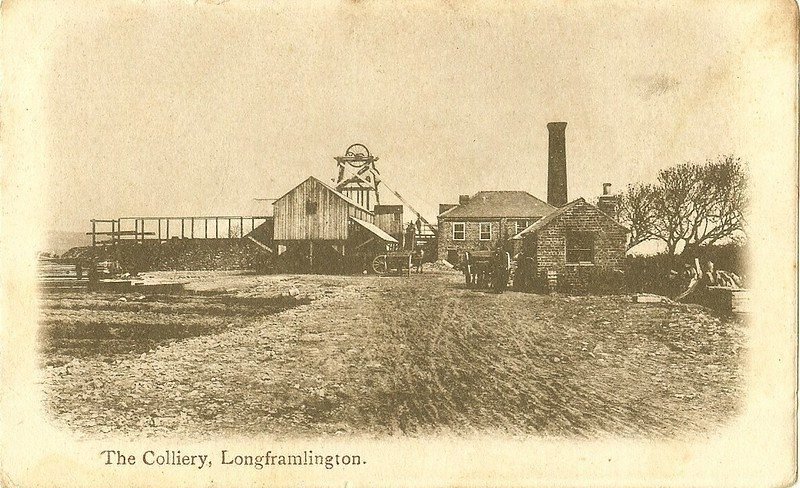 A historic photograph of The Colliery in Longframlington, featuring industrial buildings, a tall chimney, and mining structures.