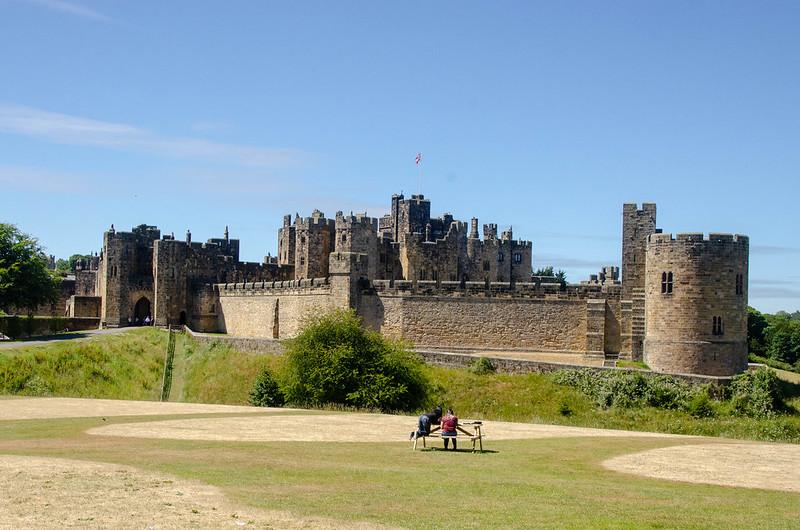 Stone castle with multiple towers and battlements, surrounded by grassy grounds and a picnic table with two people sitting on it in the foreground.