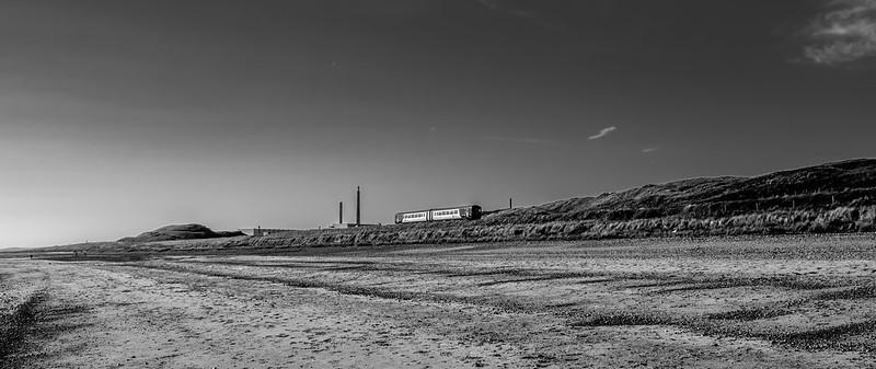 Black and white photograph of a coastal landscape with a lone train traveling, distant industrial structures, and a vast, empty beach in the foreground.