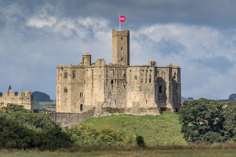 Medieval stone castle with a red flag atop the central tower, surrounded by green hills and trees under a cloudy sky.