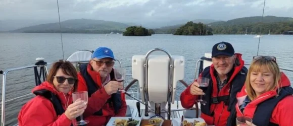 Four people wearing red jackets, sitting on a boat with drinks and food, with a lake and hills in the background.