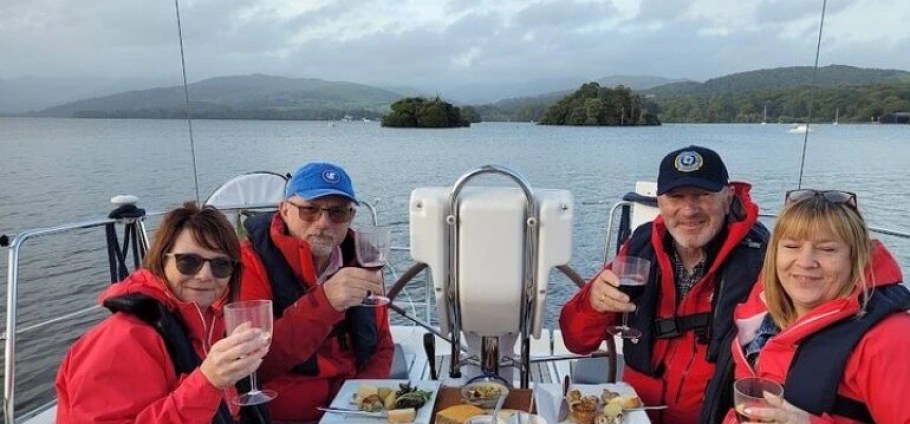 Four people wearing red jackets, sitting on a boat with drinks and food, with a lake and hills in the background.