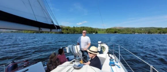 People sailing on a boat on a sunny day with a scenic view of a lake and green hills in the background.