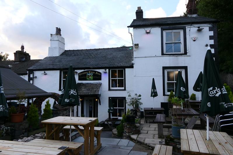 White-painted inn with grey roof, labelled "Crown & Cow Inn" on sign above entrance, outdoor wooden picnic tables, green umbrellas with "Carlsberg" branding.