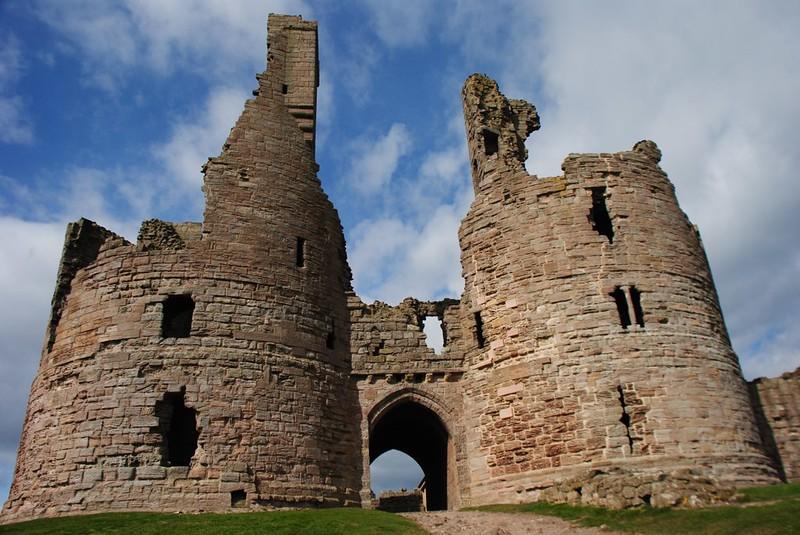 Dunstanburgh Castle, Northumberland | Ruins of a stone castle with two round towers and an arched entrance against a blue sky with clouds.