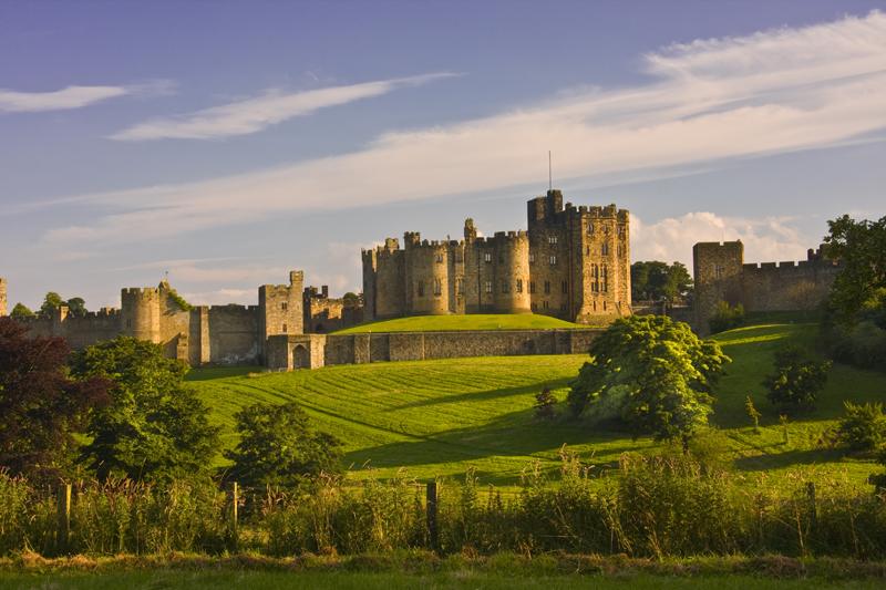 Alnwick Castle | Historic stone castle with multiple towers and crenellations, surrounded by lush green landscape and a clear blue sky.