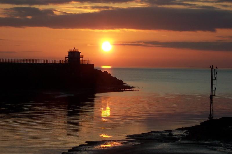 workington sunset | A scenic sunset over a calm sea with the silhouette of a lighthouse and a pier in the distance, reflecting on the water.