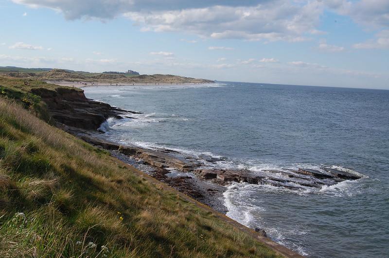 Rocky coastline with grassy cliffs, waves crashing against the shore, and a sandy beach in the distance under a partly cloudy sky.