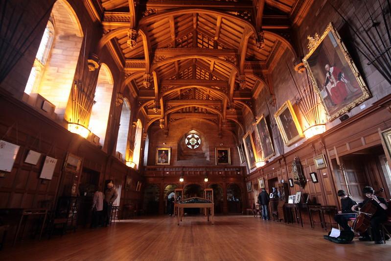 Grand hall with wooden floors, vaulted wooden ceiling, arched windows, and walls adorned with framed portraits and paintings.