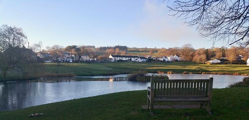 Park bench by a pond with a village and rolling hills in the background.