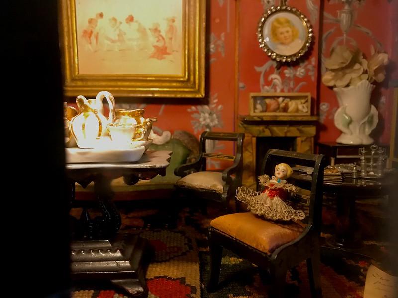 Hill top Farm Near Sawley | Detailed dollhouse room with vintage miniature furniture, a porcelain tea set on a table, and a doll on a chair. The background features floral wallpaper, framed pictures, and decorative items.