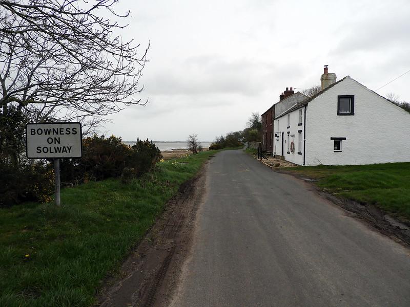 Bowness-on-Solway village sign near a narrow road with houses and greenery on a cloudy day.