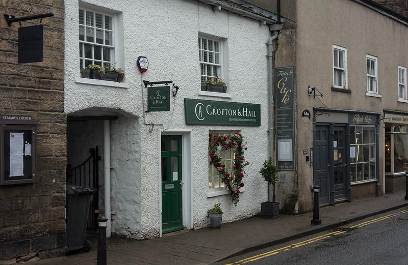 Front of a quaint street with a small shop named "Crofton & Hall," featuring a prominently displayed wreath with red roses. The shop has a white facade, a green door, and windows with flower boxes. Adjacent is a grey building housing "Tata's Café."