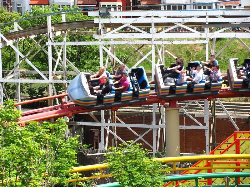 People riding a roller coaster at an amusement park with white wooden supports and greenery in the foreground.