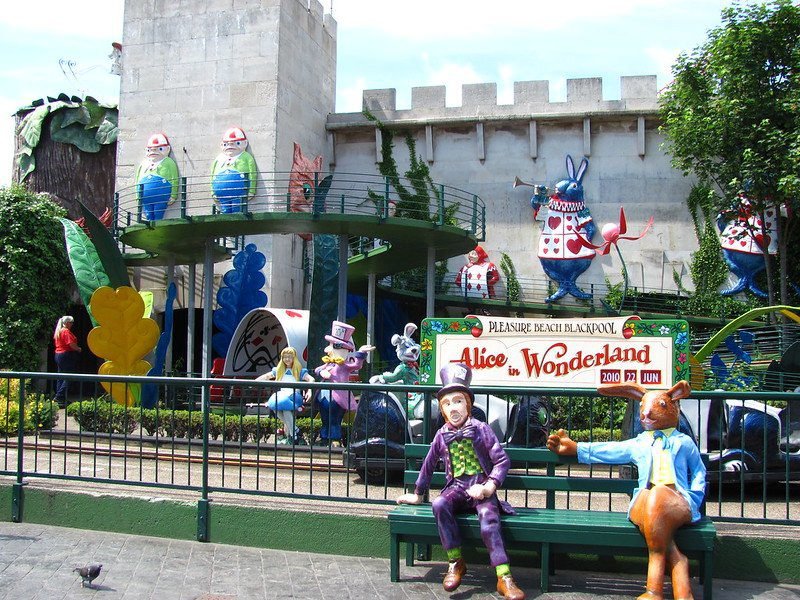 Amusement park ride themed after Alice in Wonderland, featuring colourful character statues, a castle-like building, and vibrant foliage.