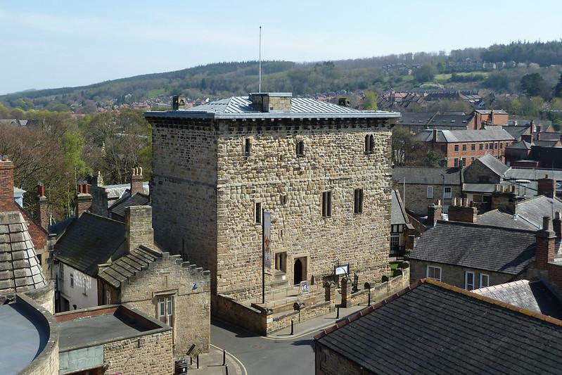 Aerial view of a historic stone building with a flagpole on top, situated in a town with houses and trees in the background.