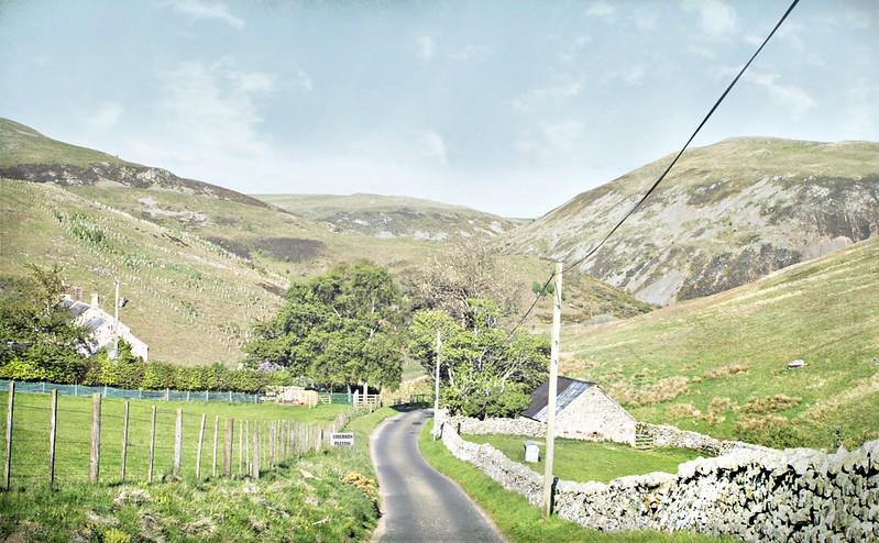 Narrow road winding through a rural valley with green fields, stone fences, scattered trees, and hills in the background.