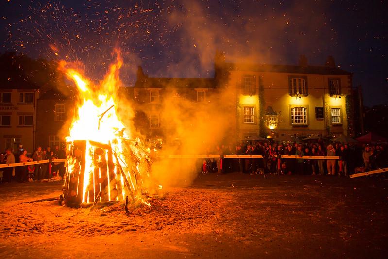 Large outdoor bonfire at night with a crowd of people watching in the background with illuminated buildings.