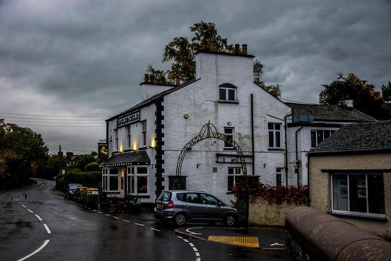 A white, two-storey building with black accents and a sign reading "THE BLACK BULL" on its facade, located by a wet, curving road on a cloudy day. A small silver car is parked beside the building.