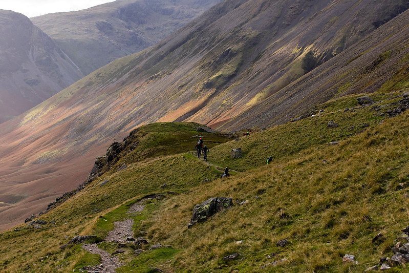 Mountain bikers on a narrow grassy trail with a steep hillside and expansive valley in the background.