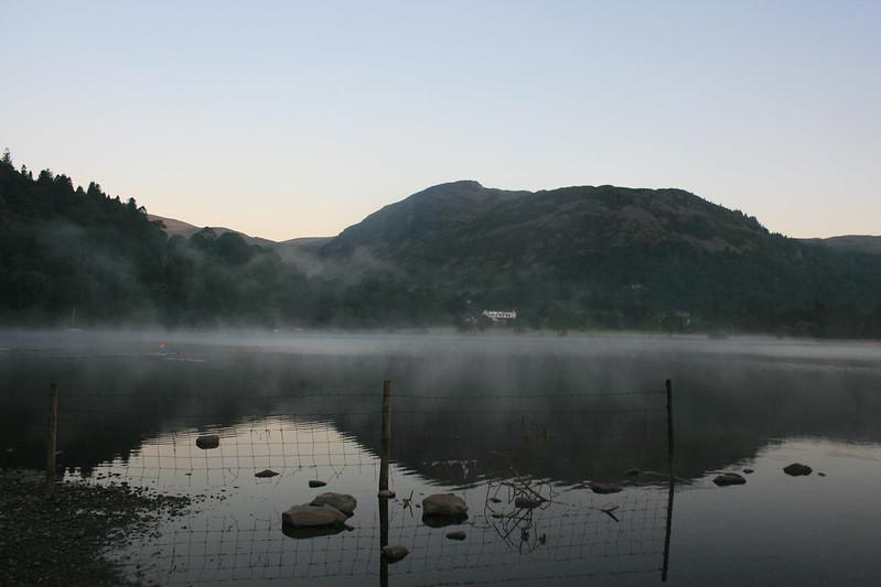 Ullswater | Mountain landscape with a misty lake, reflecting the surrounding hills and trees at dawn, and a wire fence in the foreground.