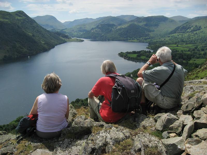 Three people sitting on a rocky hilltop, looking out over a large lake surrounded by green mountains and forests.