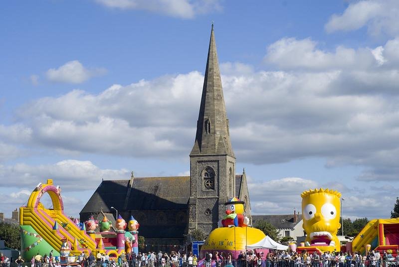 Silloth, Cumbria | A Crowd of people at a funfair with large inflatable attractions in front of a church with a tall spire, under a partly cloudy sky.