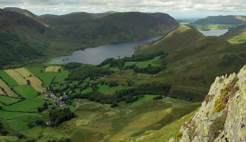 Buttermere Village, Cumbria | Aerial view of a lush green valley with farm fields, a small village, a lake, and surrounding hills.