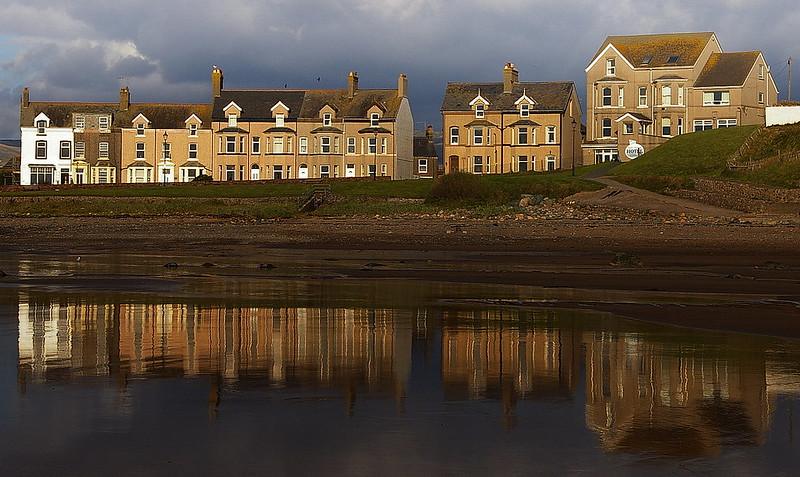 Row of old terraced houses and a larger house reflected in a body of water with a cloudy sky above.