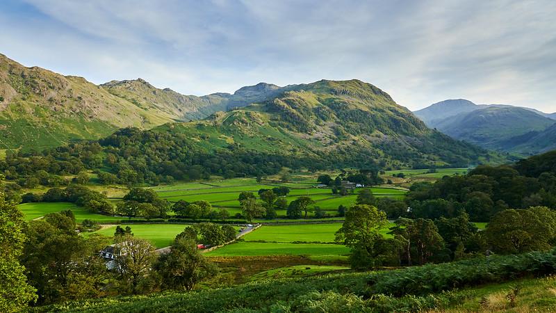 A picturesque landscape of verdant fields, dense woodlands, and rolling hills under a clear sky.