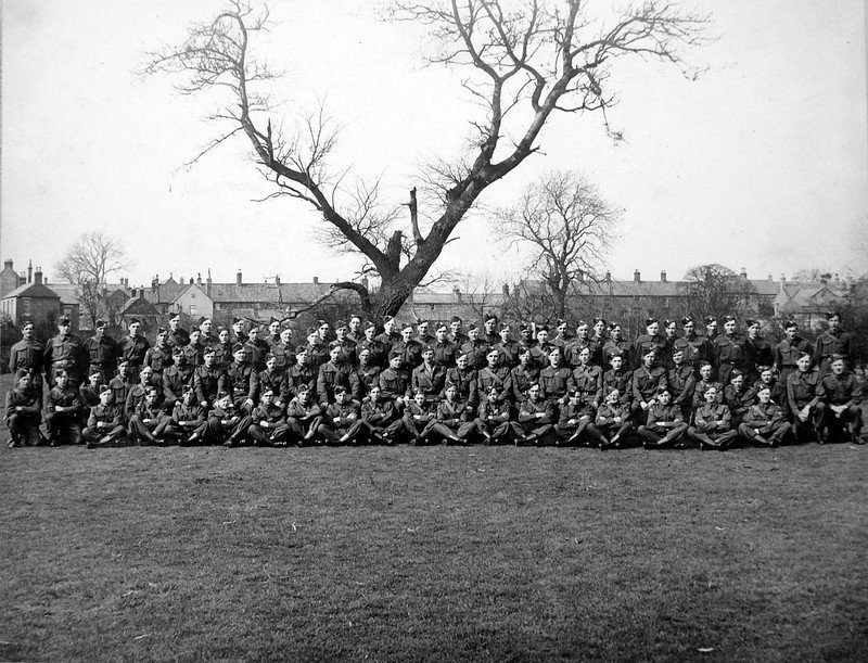 A large group of soldiers in uniform are sitting and standing in rows on a grassy field in front of a leafless tree, with houses visible in the background.