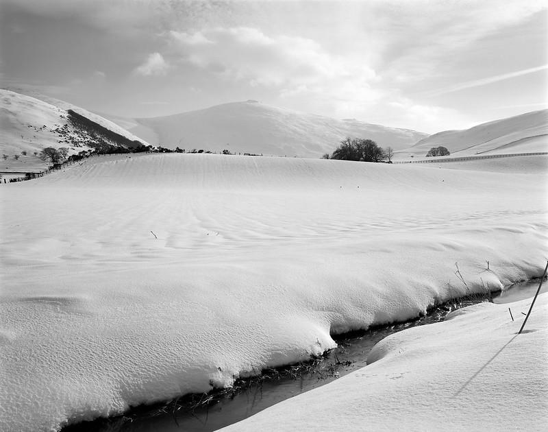 Snow-covered landscape with a small stream, hills, and trees in the distance under a partly cloudy sky.