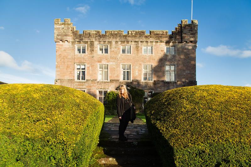 A person standing between manicured hedges in front of a historic stone building under a blue sky.