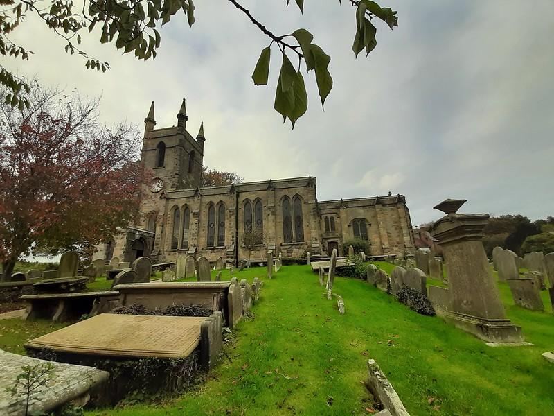 Old church with tall arched windows, a clock tower, and surrounding gravestones on a grassy hill.