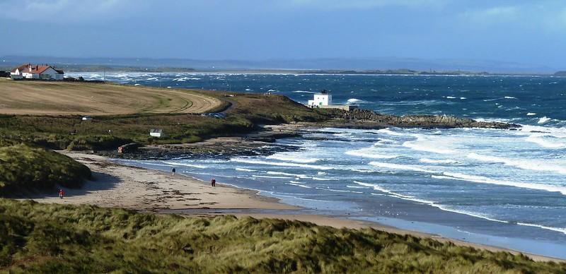 Sandy beach with grassy dunes, a few people walking, a white house near the sea, and a field with a house in the background under a cloudy sky.