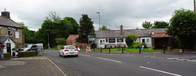 A road junction in a village featuring a white car, road signs, and various buildings including a pub and shops with green trees in the background.