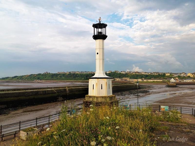 Maryport Lighthouse | A white lighthouse with a black top stands on a grassy area near the coast, with buildings and a bay visible in the background under a cloudy sky.