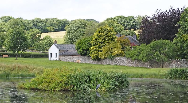 Countryside scene with a white house, stone wall, and lush green trees by a pond.