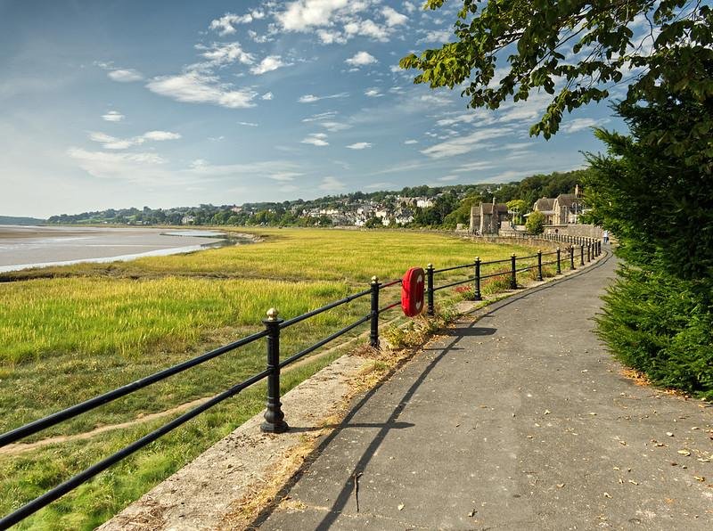 Grange Over Sands Promenade | A paved pathway with a black railing runs alongside a grassy landscape and an estuary, with a lifebuoy attached to the railing. Houses and buildings in the distance are nestled among green trees under a partly cloudy sky.