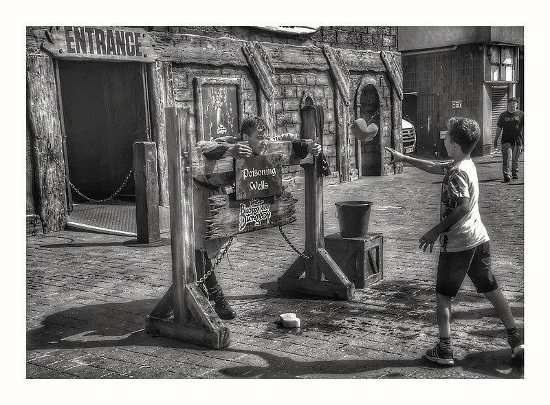 A child throws a soft object at another child in stocks labeled "Poisoning Wells" outside a rustic-themed building entrance.