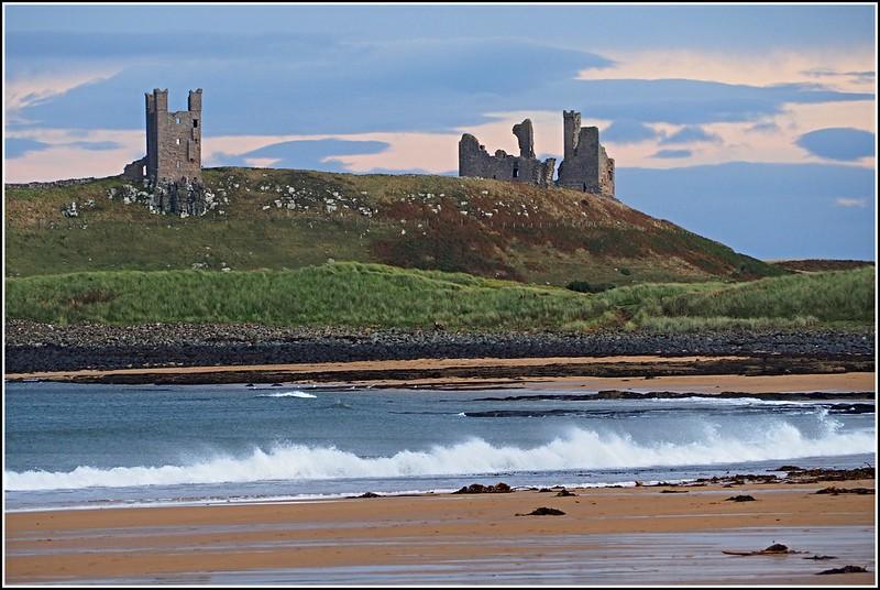 Ruined castle on a grassy hill overlooking a sandy beach with waves crashing on the shore.